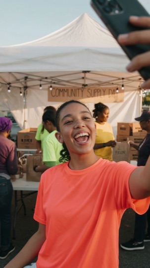A volunteer in an orange shirt filming herself at a community supply drive, with other volunteers packing boxes under a tent behind her.